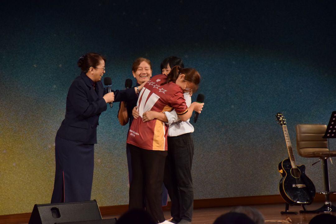 黎欣慧和妈妈叶玉英在新春发放的舞台上相拥，志工黄舒莉（左二）欣喜见证两母女经过风雨考验，终迎来温暖的骄阳。【摄影：李莉芬】